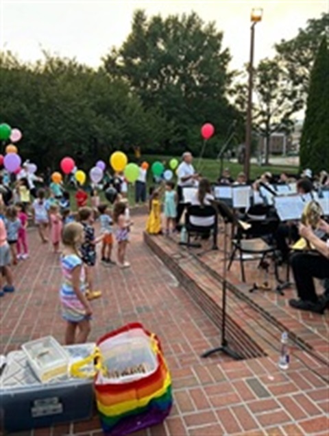 Young children holding balloons watching a small band performance on a brick stage