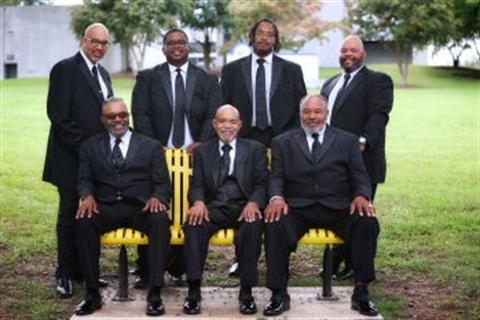 A group of 8 men, 4 standing up behind 4 sitting on a yellow bench. All dressed in black with white shirt and dark tie. Smiling facing the camera. 