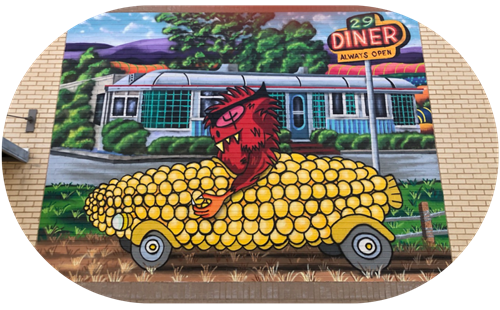 Mural of a red creature driving a corn cob car in front of 29 Diner, with the sign reading “Always Open.”