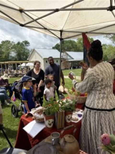 A woman in Civil War clothing behind a table with historical objects talking to a family with three children. In the background is the Historic Blenheim Civil War Interpretive Center.