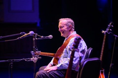 Man sitting down and facing left with microphones in front of him and a guitar perched behind him, purple lighting effect.